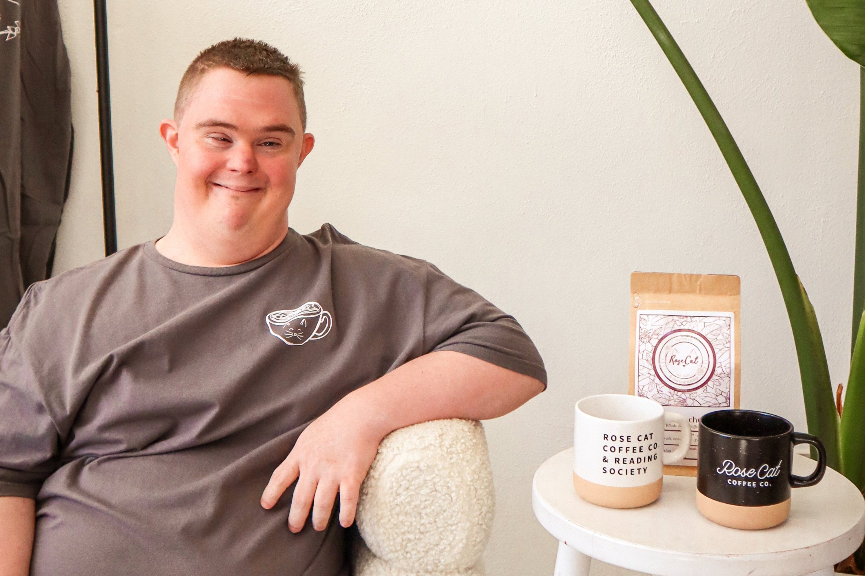 Rose Cat Coffee Co Barista, Chad, posing in a tshirt next to two Rose Cat Coffee Mugs and Rose Cat Coffee beans, available in store or online. 